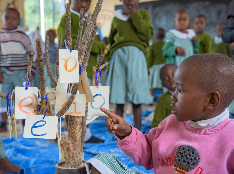 child pointing to painted letters on pieces of paper hanging from a branch inside a classroom in tanzania